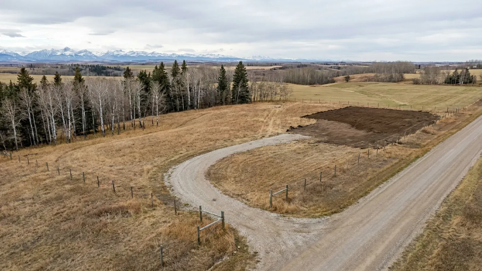 Aerial view of an undeveloped acreage of rolling grassland with an aspen stand and a gravel approach, hay fields and distant snow-capped mountains beyond.
